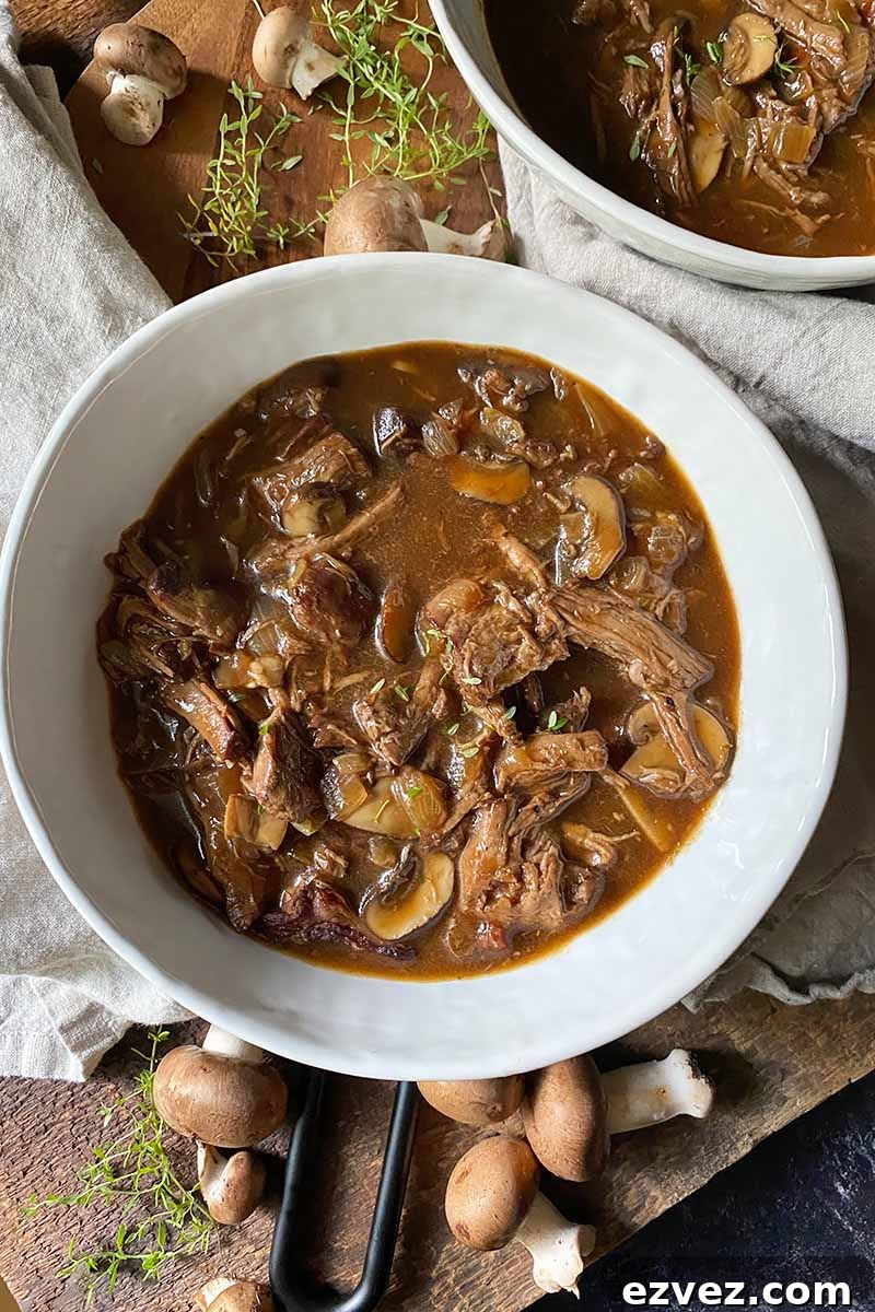 Leftover Pot Roast Mushroom Soup Recipe for Comforting Meals 3 Vertical top-down image of two white bowls filled with a meat stew surrounded by herbs and mushrooms.
