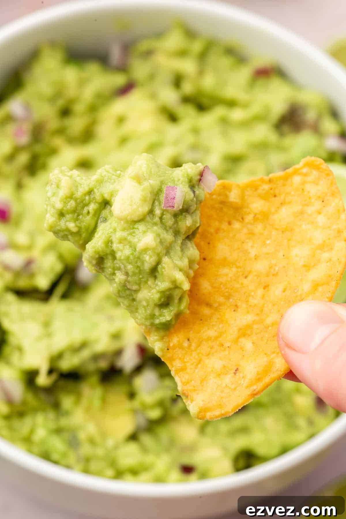 Quick 4-Ingredient Guacamole Recipe 4 A hand holding a tortilla chip dipped in guacamole over a bowl of more guacamole.