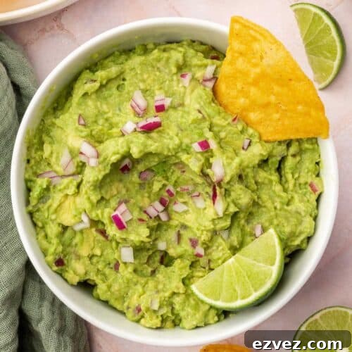 Quick 4-Ingredient Guacamole Recipe 5 An overhead view of a bowl of guacamole topped with diced red onion, a tortilla chip and a lime wedge with additional chips and limes on the surface.
