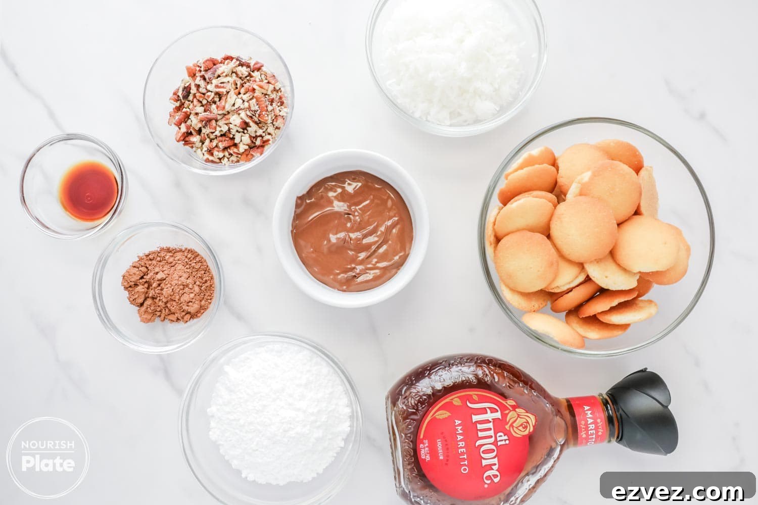 Overhead view of Amaretto ball ingredients arranged in glass bowls on a white surface, including vanilla wafers, chopped pecans, shredded coconut, powdered sugar, cocoa powder, Nutella, pure vanilla extract, and a bottle of Amaretto liqueur.